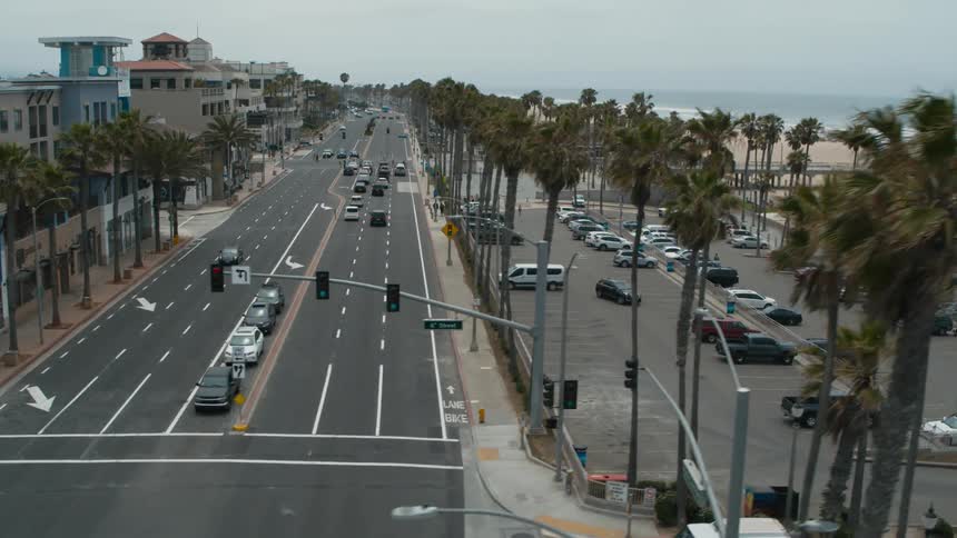 An aerial view of cars driving on a street and in a parking lot by a beach with stores across the street and people walking around