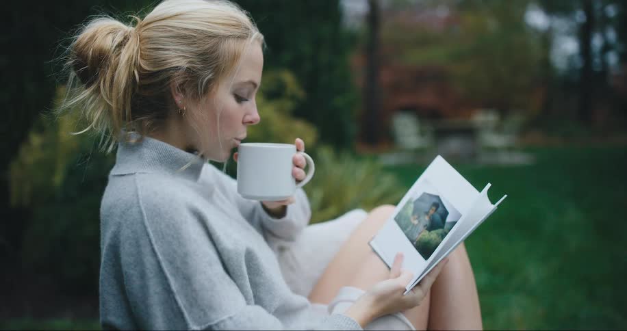 Woman sits in yard reading and drinking coffee