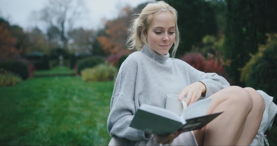 A woman sits in her yard reading a book and drinking coffee