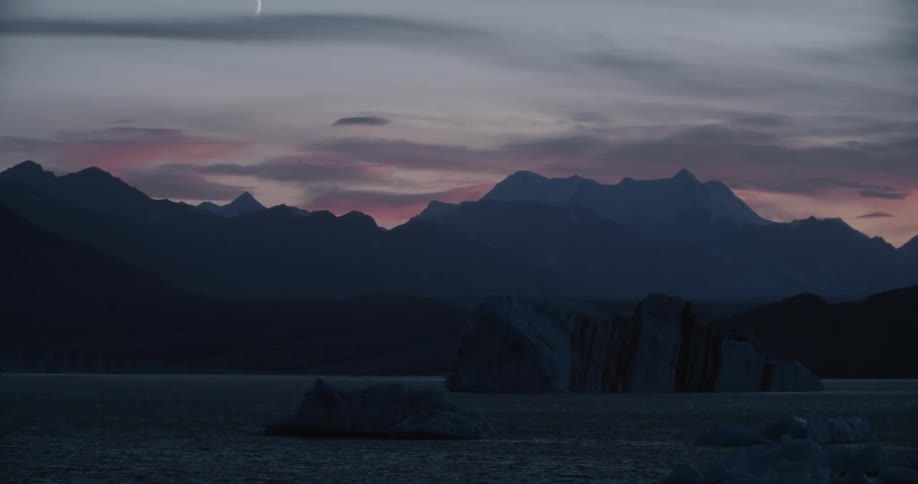 Icebergs are in the water with mountains in the distance and a crescent moon in a colorful sky