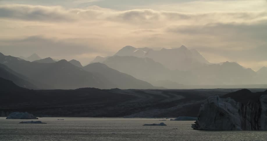 Mountain ridges above waves splashing against a rocky shore under a cloudy sky