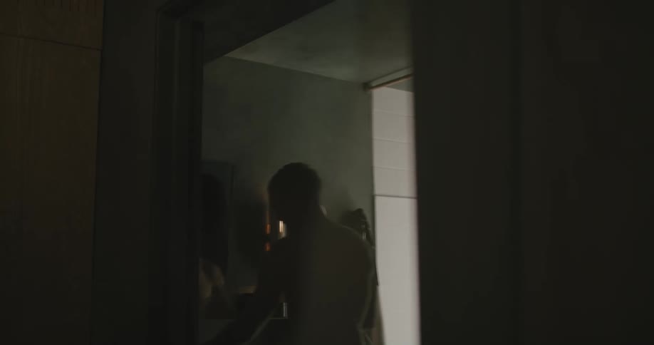 A man walks up to a bathroom mirror and wipes his head with a towel