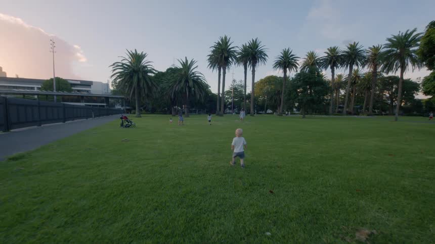 A young child in a park walks towards a man playing badminton with his children