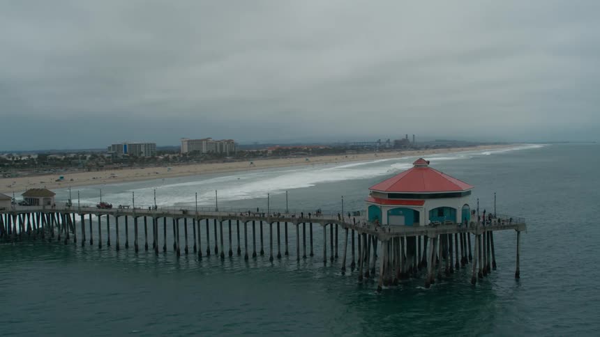 Waves roll over a beach near a coastal community as people walk around a building at the end of a pier