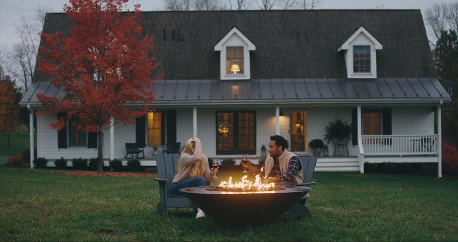 Couple sits in front of their house by a fire pit drinking wine