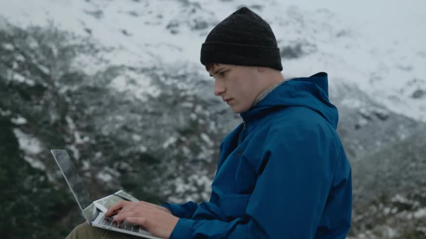 A man wearing a coat and a knit cap is sitting outside near snow covered mountains typing on a laptop computer