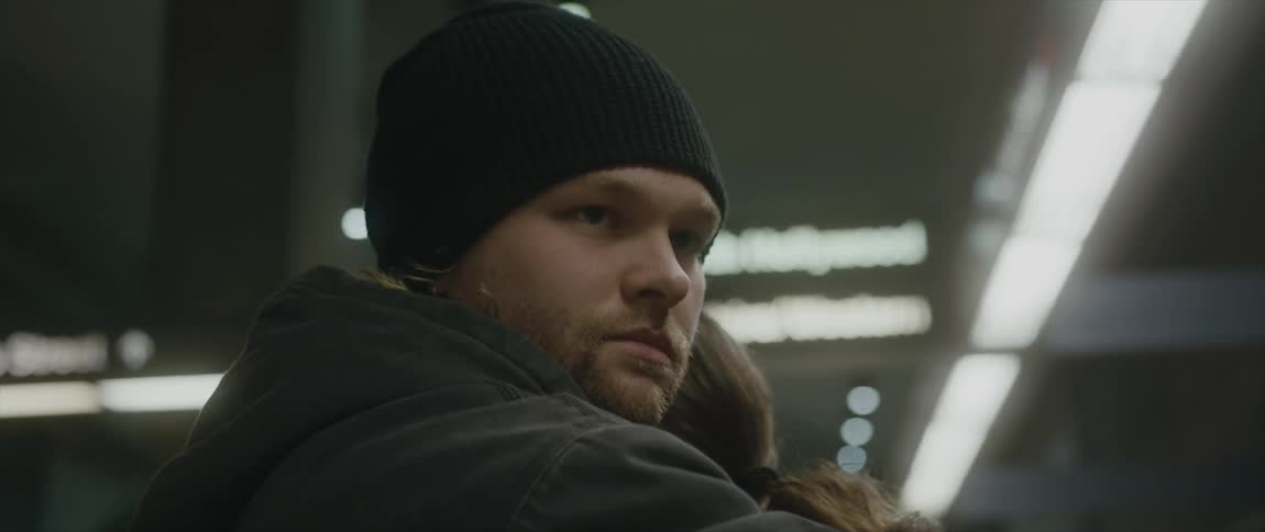 A man wearing a coat and a knit cap is hugging a woman under bright lights