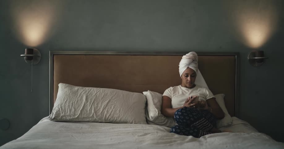 A woman with a towel around her head is sitting on a bed and using her phone