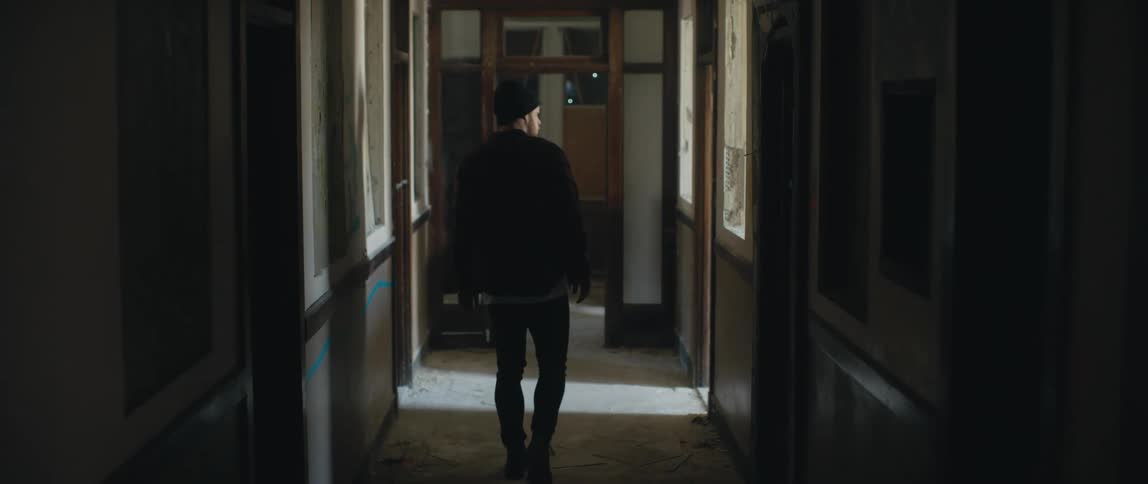 A man wearing a flannel shirt and a knit cap is walking down the hallway of an apartment building looking at doors in a suspicious manner