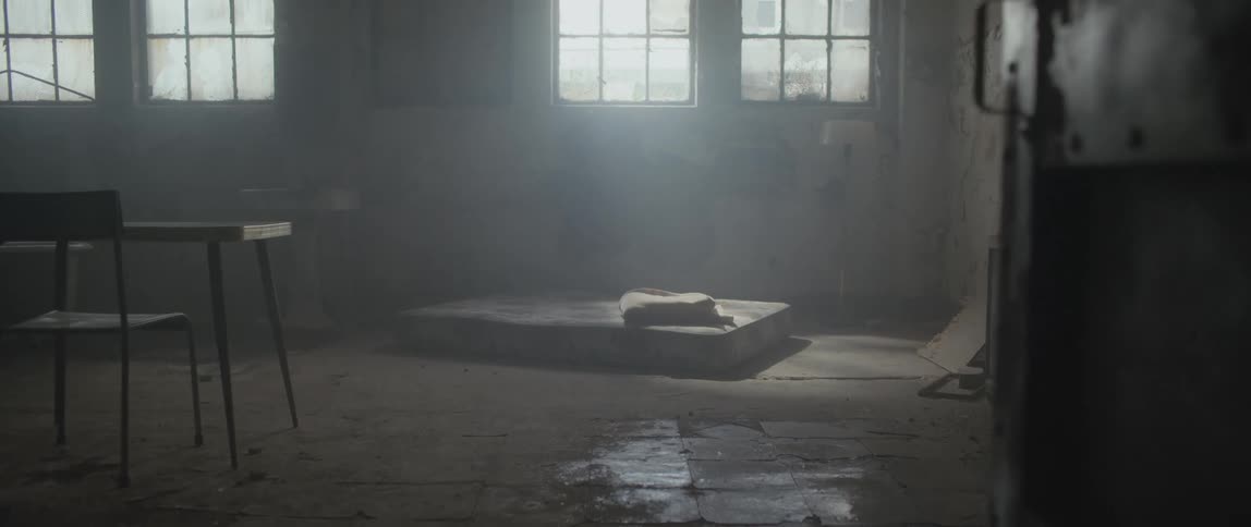 A table with a chair and a mattress are on the floor of a dilapidated room