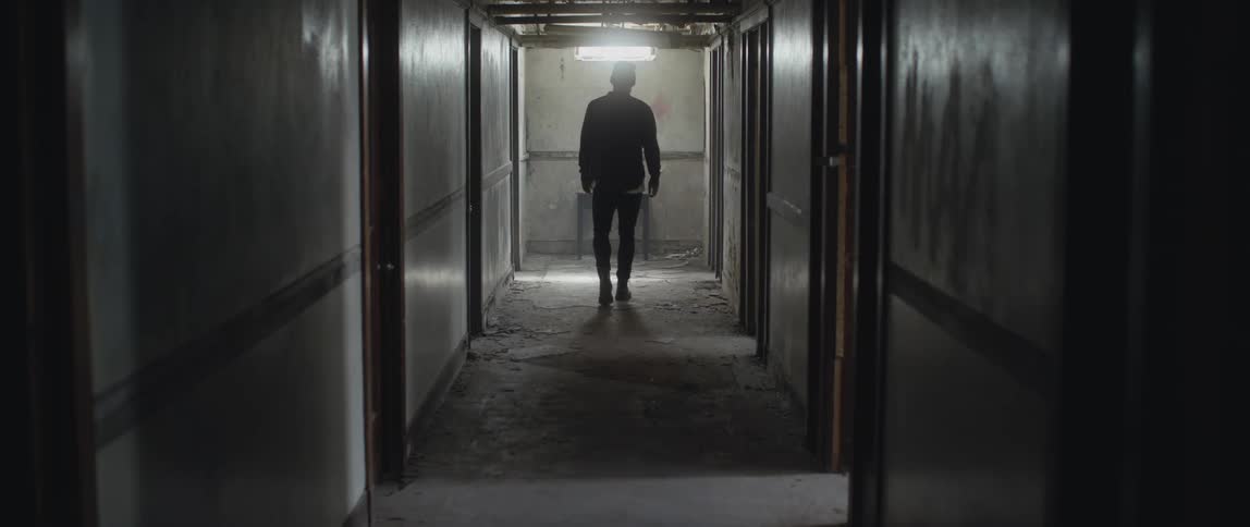 A man wearing a flannel shirt and a knit cap is walking down the hallway of a dilapidated building