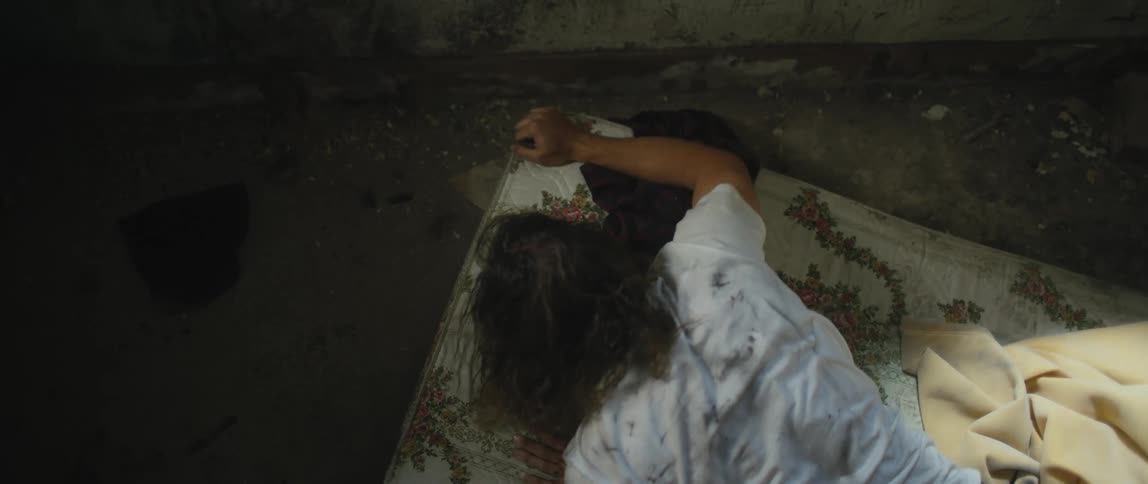 A man covered in dirt lies face down on a mattress in a filthy room and attempts to get up and picks up a stone and puts a mark on the wall