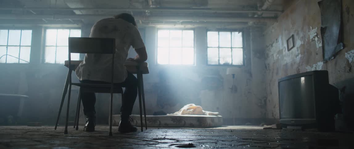 Man sits on a chair at a small table in a room that has a mattress on the floor and an older television against a wall