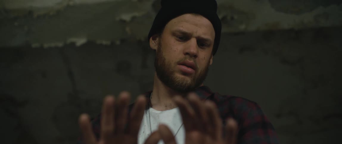 A man wearing a flannel shirt and a knit cap is looking at his hands and appears to be disoriented and then turns away