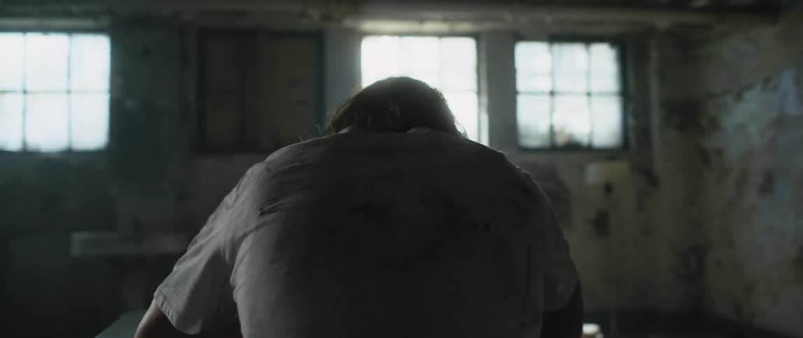 Man wearing dirty shirt sits hunched over table in room with peeling paint