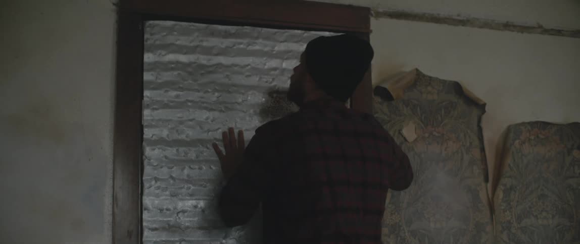 A man wearing a longsleeved heavy work shirt and a black beanie runs into a closing door as it slams shut pushes against it and then walks back into the room shaking his head