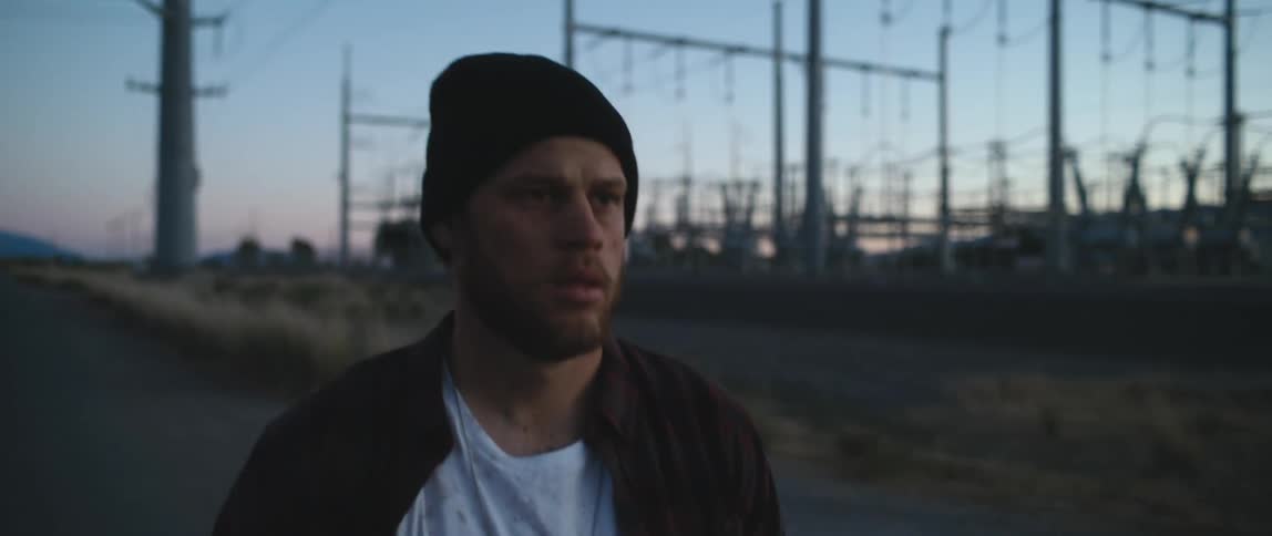 Young bearded man wearing a beanie walking down a rural road near a power substation