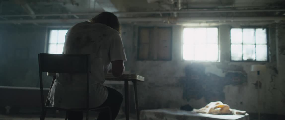 A man is sitting at a table in an old abandoned building and he stands up and walks