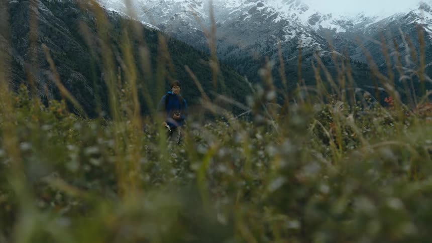 A man walks up a slope of green grass and plants with snow dusted mountains behind him holding a notebook and camera