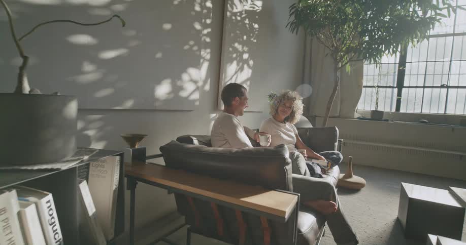Woman with curly hair t shirt pants sits on couch next to man wearing white sweatshirt and jeans holding a cup in living room at home
