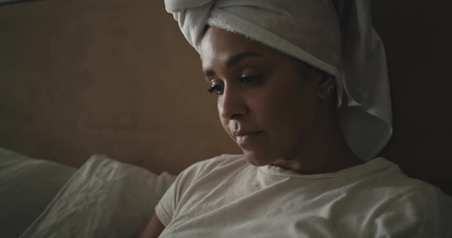 Woman reclines in bed against headboard with towel wrapped over her hair