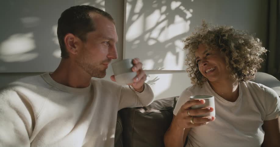 A man and a woman are sitting on a couch talking and laughing as they drink from cups