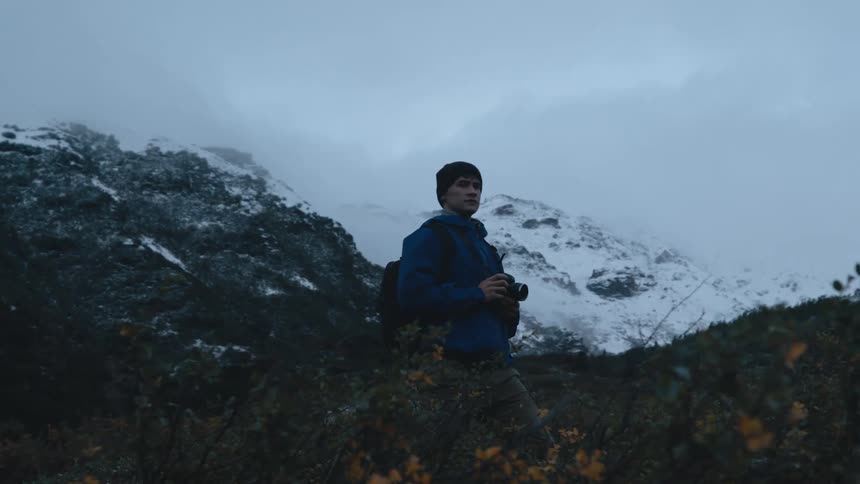 A man carrying a camera hikes through a mountain range and stops and looks around