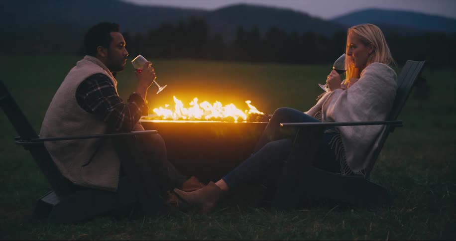Couple sits outside near a fire drinking wine