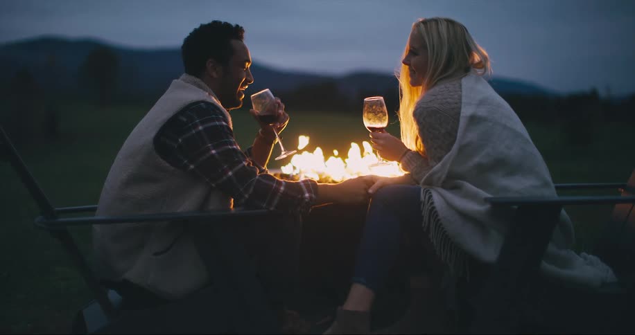 Couple sits outside by a fire with wine