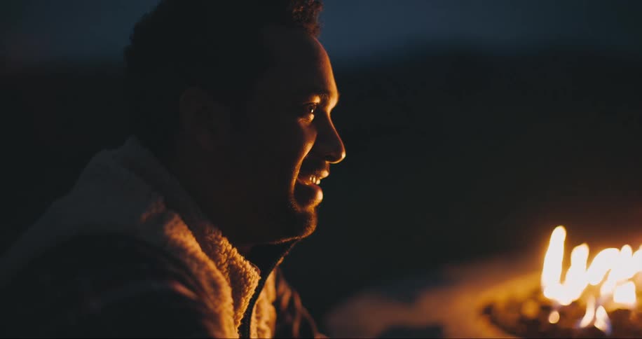 Man sits by campfire smiling outdoors at night