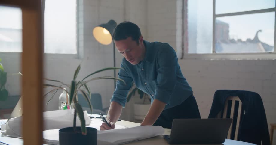 Man with blue shirt and dark blue pants writes down in notebook looking at laptop in the office