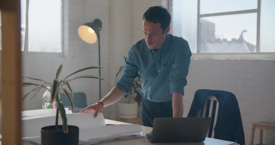 A man in an office is standing and leaning over his laptop and large blueprint papers during the day