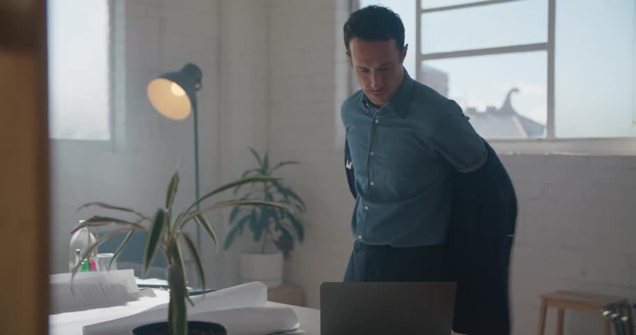 A man enters his office and opens his laptop before removing his jacket and taking a seat at his desk