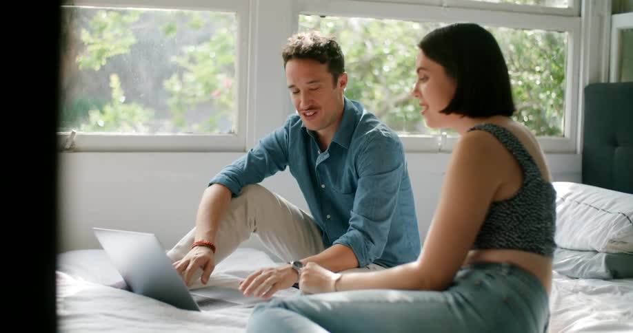 A man and a woman sitting on a bed and talking while the man types on a laptop