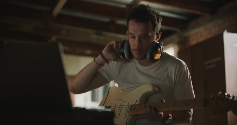 A man playing guitar pauses and takes his headphones off to make adjustments on a keyboard