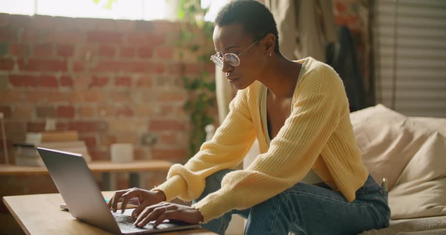 A woman looks at a notepad while typing on her laptop while sitting on the couch