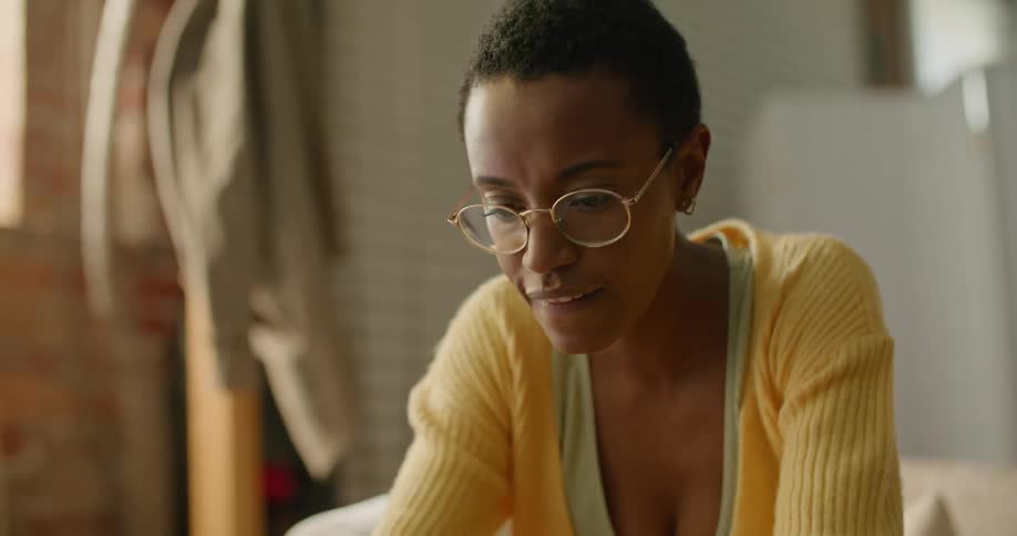 Woman with short hair glasses and a yellow sweater sits on couch and works on laptop in the living room at home