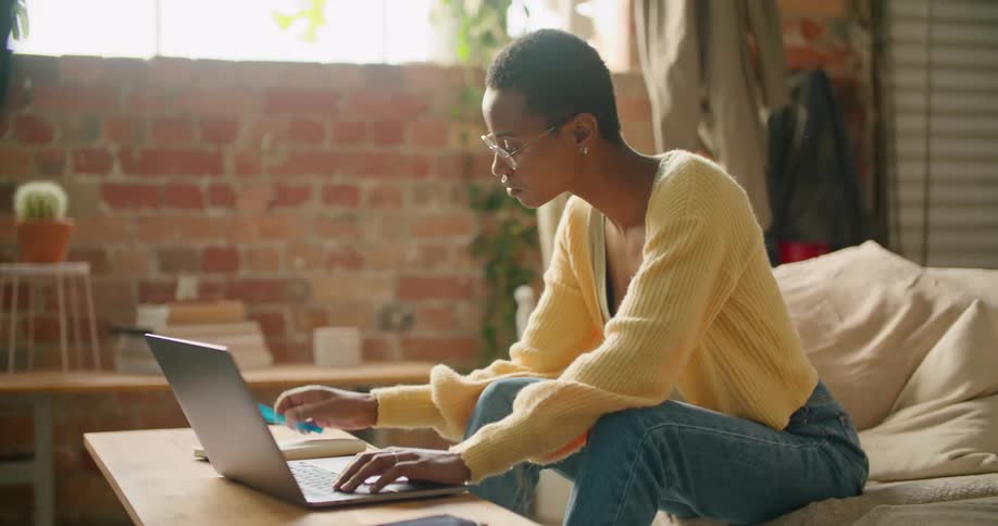 A woman in a room sitting on a couch typing on a laptop with a wall in the background