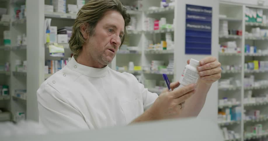 A pharmacist reads notes and then checks a pill bottle