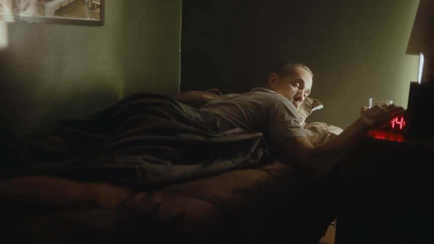 A man tiredly reaches for the alarm clock turns it off and squints into the daylight before sitting up