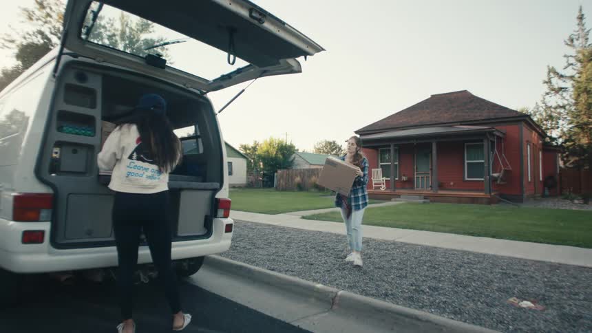Two women load rear of van parked at curb in front of house
