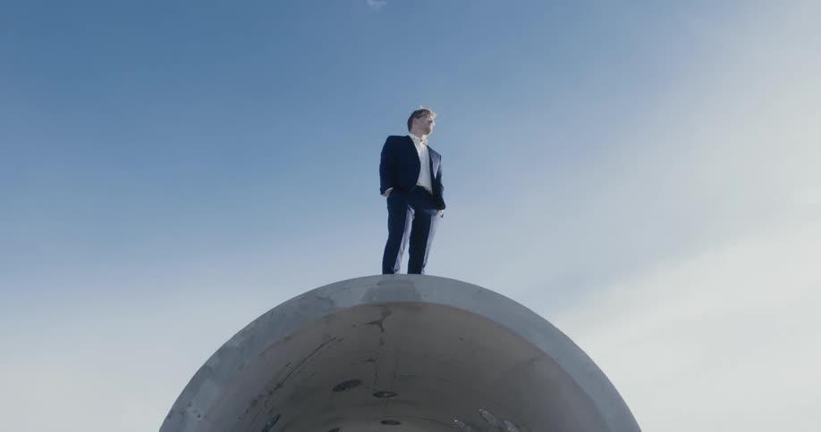 Man in business suit stands on top of large concrete segment of tunnel in a desert backed with mountains