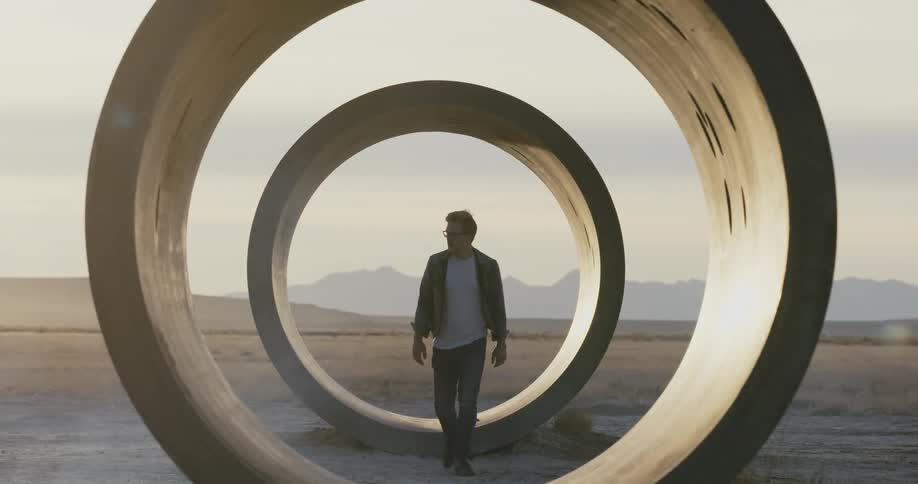 A man walks through large sewer pipes that are sitting in an open field and there are mountains in the distance