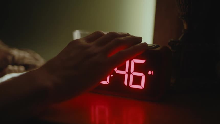An alarm clock is on a table and someone reaches out of bed and slams their hand on top of it