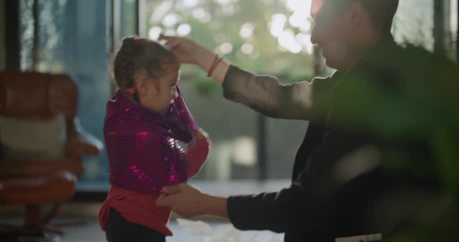 A father is in a living room helping his little girl put on a sequined jacket