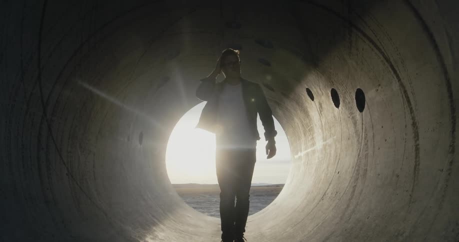 A man walks through a section of pipe with the sunshine glaring behind him