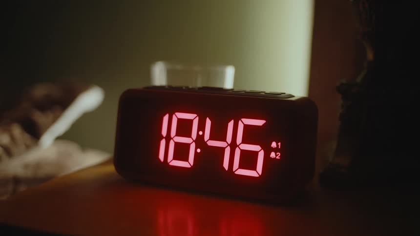 A person in a bed reaches over to press a button atop a digital alarm clock on a nightstand