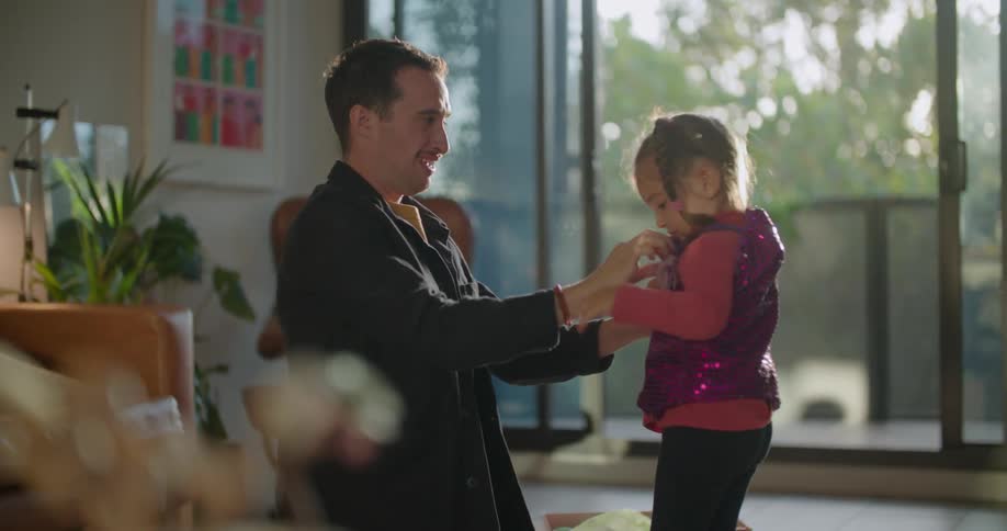 A father is in a living room helping his little girl put on a sequined jacket