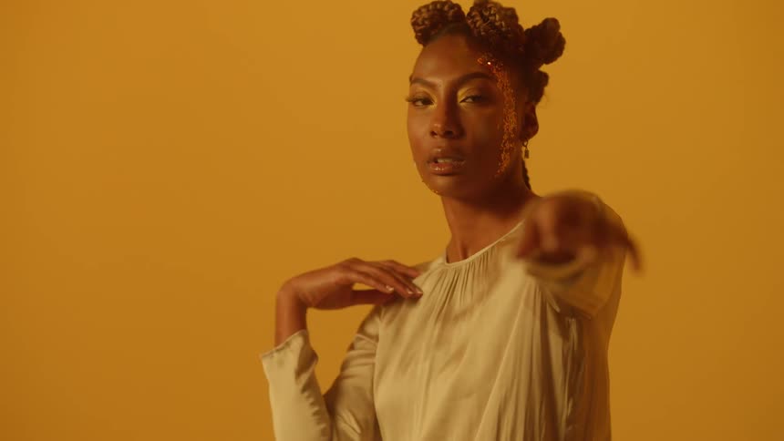 A black woman with glitter on her face is dancing near a bright yellow wall