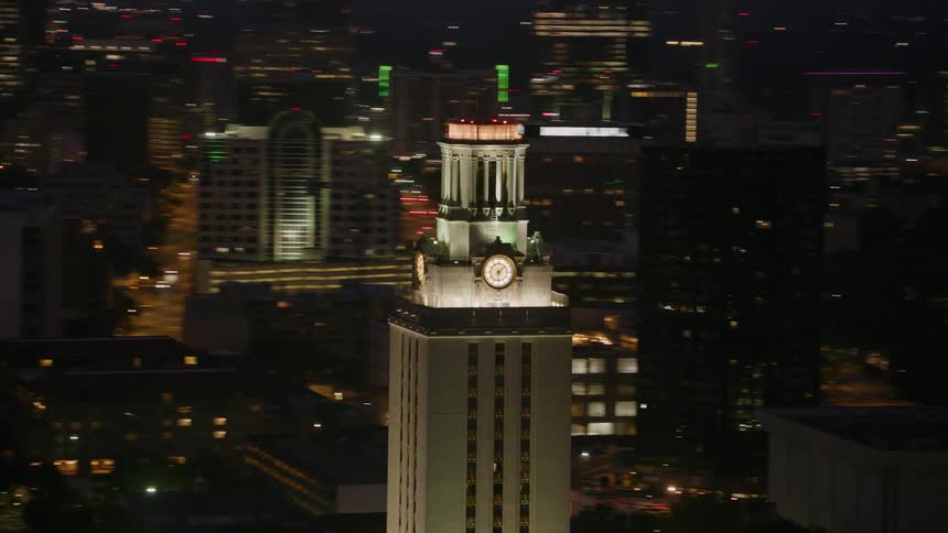 Lights are on a clocktower at night and lights on other downtown skyscrapers and buildings glow in the night sky